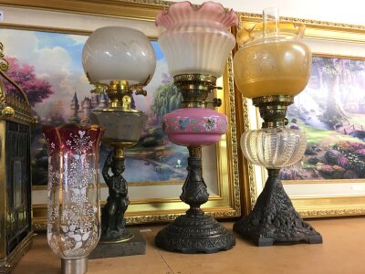 Three oil lamps featuring embossed yellow glass, etched glass shade figure lamp, and pink hand-painted reservoirs and a glass shade with hand painted white floral patterned decoration