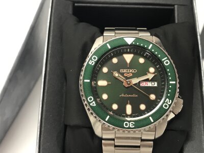Seiko 5 Sports features a green dial and matching bezel paired with a stainless steel bracelet. Seen running. Box and card.