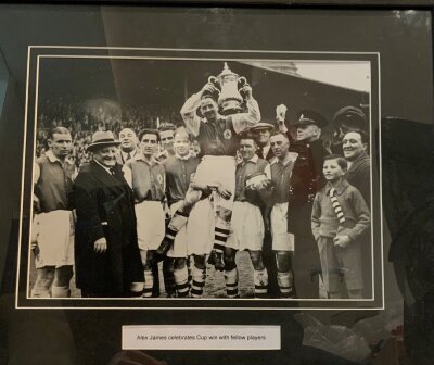 1936 Arsenal FA Cup Winning Celebration Photo: Depicts Alex James holding the cup aloft. This was part of the Highbury Stadium Sale. Comes with COA and measures approximately 55 x 44cm. Slightly scratched to the frame.