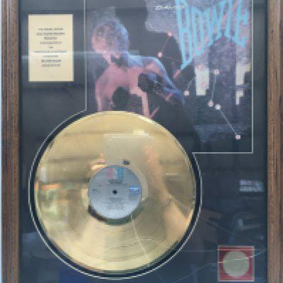 A framed David Bowie gold disc presented in recognition of a million sales of the 'Lets Dance' LP, approx 52.5cm x 68cm. Glass needs replacing.