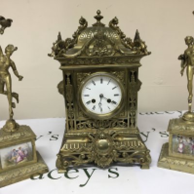 A late 19th Century brass cased clock garniture the elaborate clock case with raised scrolls. The side ornaments with figures of Mercury and set with hand painted porcelain panels. decorated with figures. The movement striking on a bell and with pendulum.